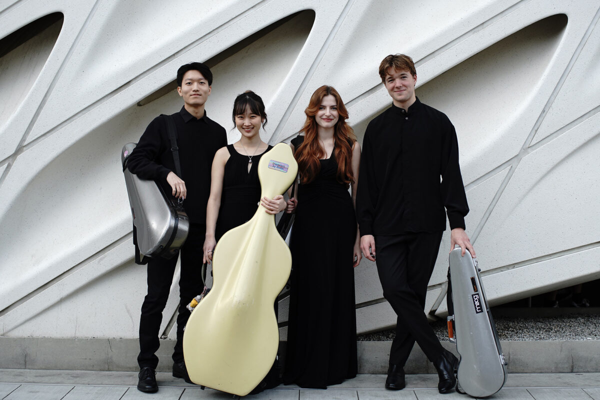 Photo of the Odyssey String Quartet posing with their instruments in front of a modern, sculptural white wall.