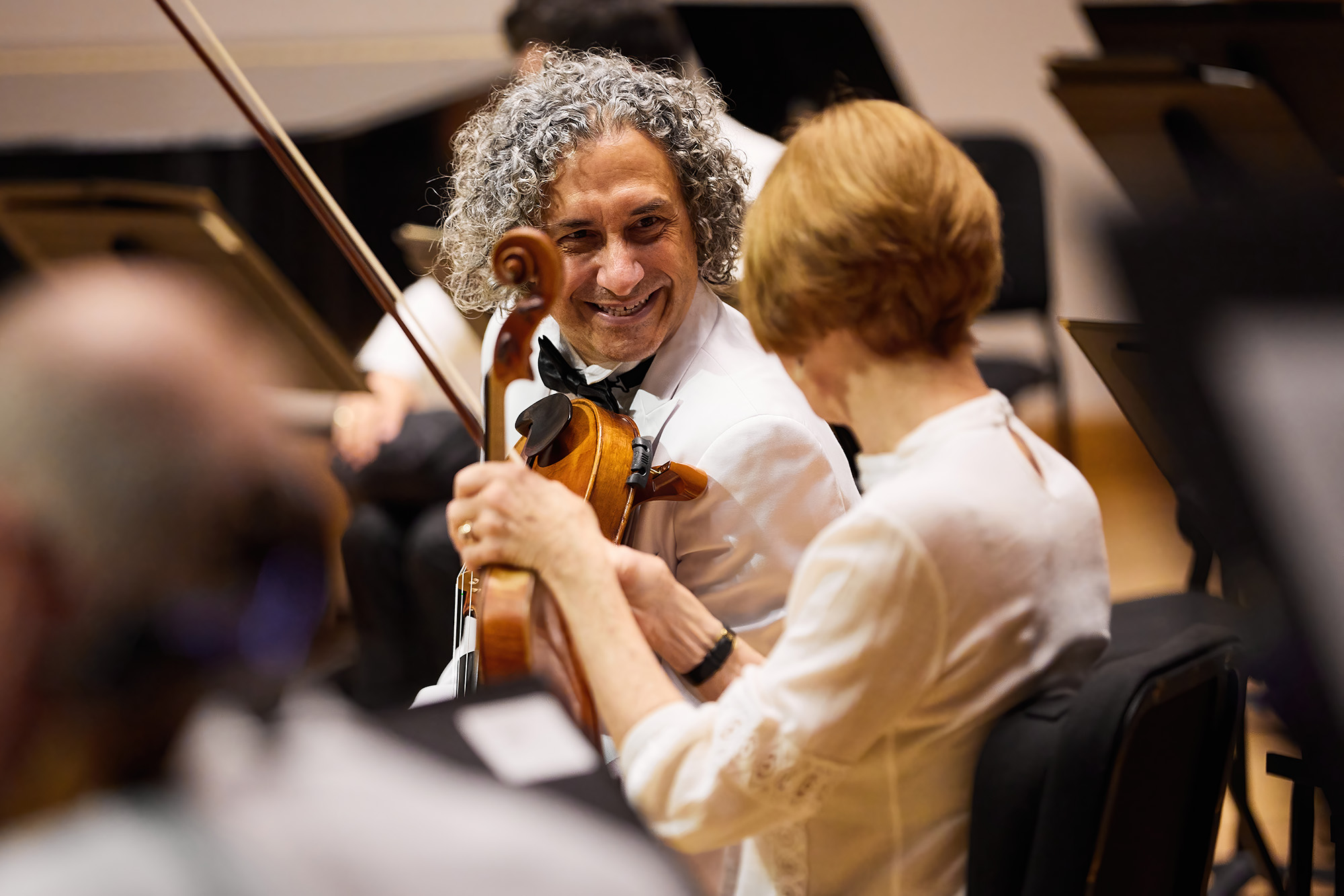 Photo of a smiling orchestra member with a violin, looking at the musician seated next to him.