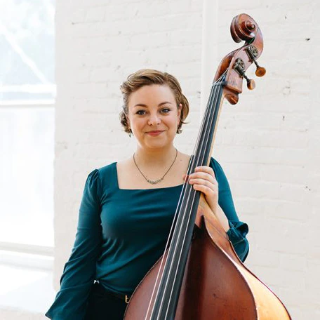 Portrait of Nina DeCesare, bass, standing in front of a white brick wall and holding a stand up bass.