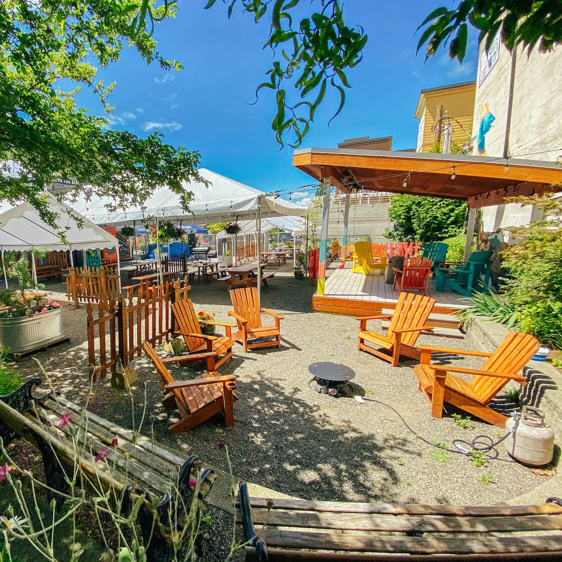 Sunny photo of the beer garden at Boundary Bay Brewing, showing a stage, white tents and adirondack chairs circling a fire pit.