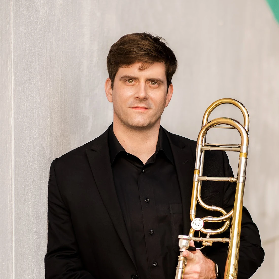 Portrait of Ross Holcombe, trombone in a black shirt, leaning against a wall and holding a trombone.