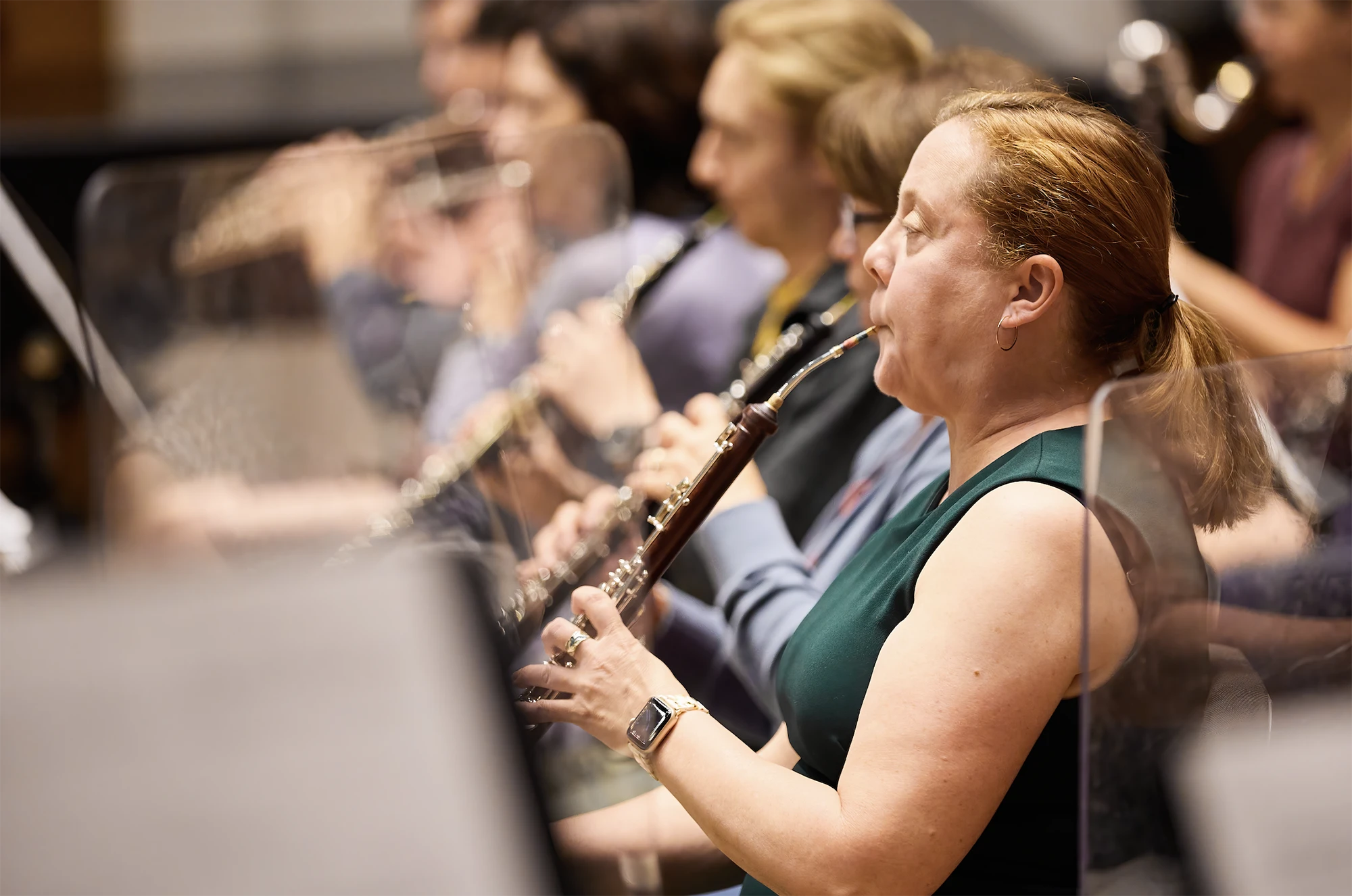 Female oboe player performing with a woodwind section during a live orchestra rehearsal, musicians playing clarinets and flutes in background, classical concert setting.