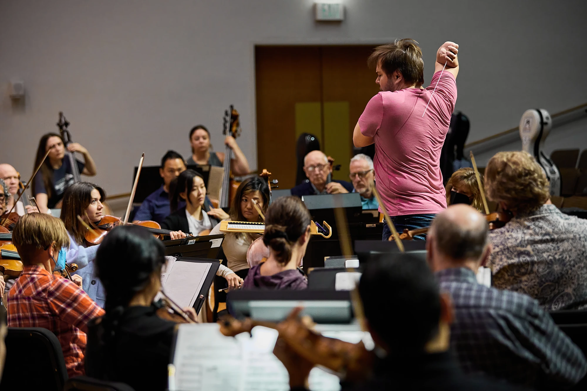Conductor Marcelo Lehninger leads a symphony orchestra rehearsal, passionately directing string musicians with his baton in a concert hall setting.