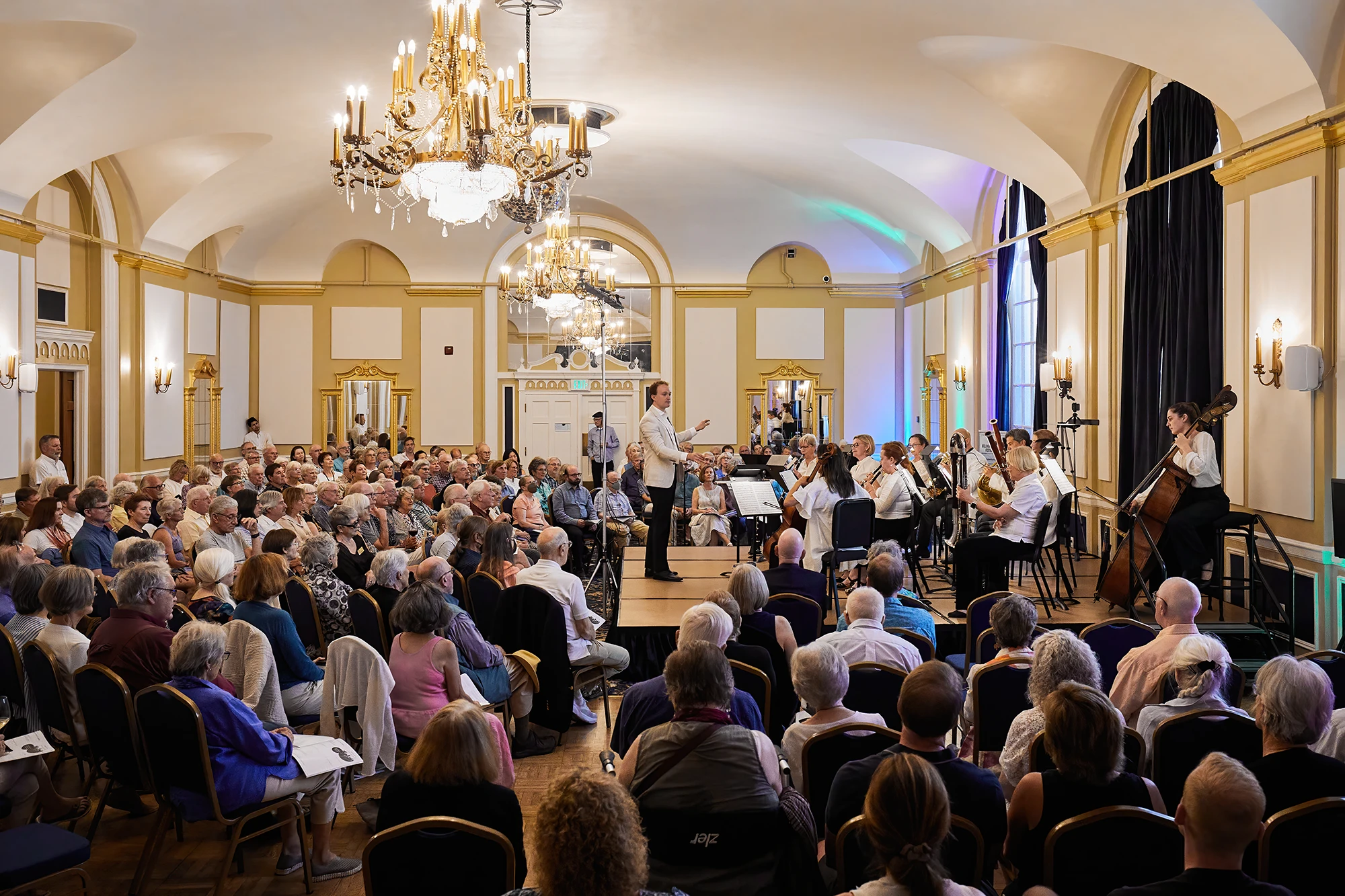 Conductor Ryan Dakota Farris leads a chamber orchestra performance in an elegant concert hall with chandeliers, as a large audience enjoys the live classical music event.