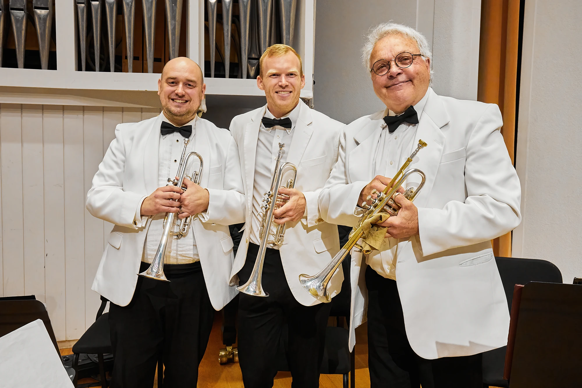 Three trumpet players in formal white tuxedos with black bow ties posing backstage with their instruments before an orchestra performance.