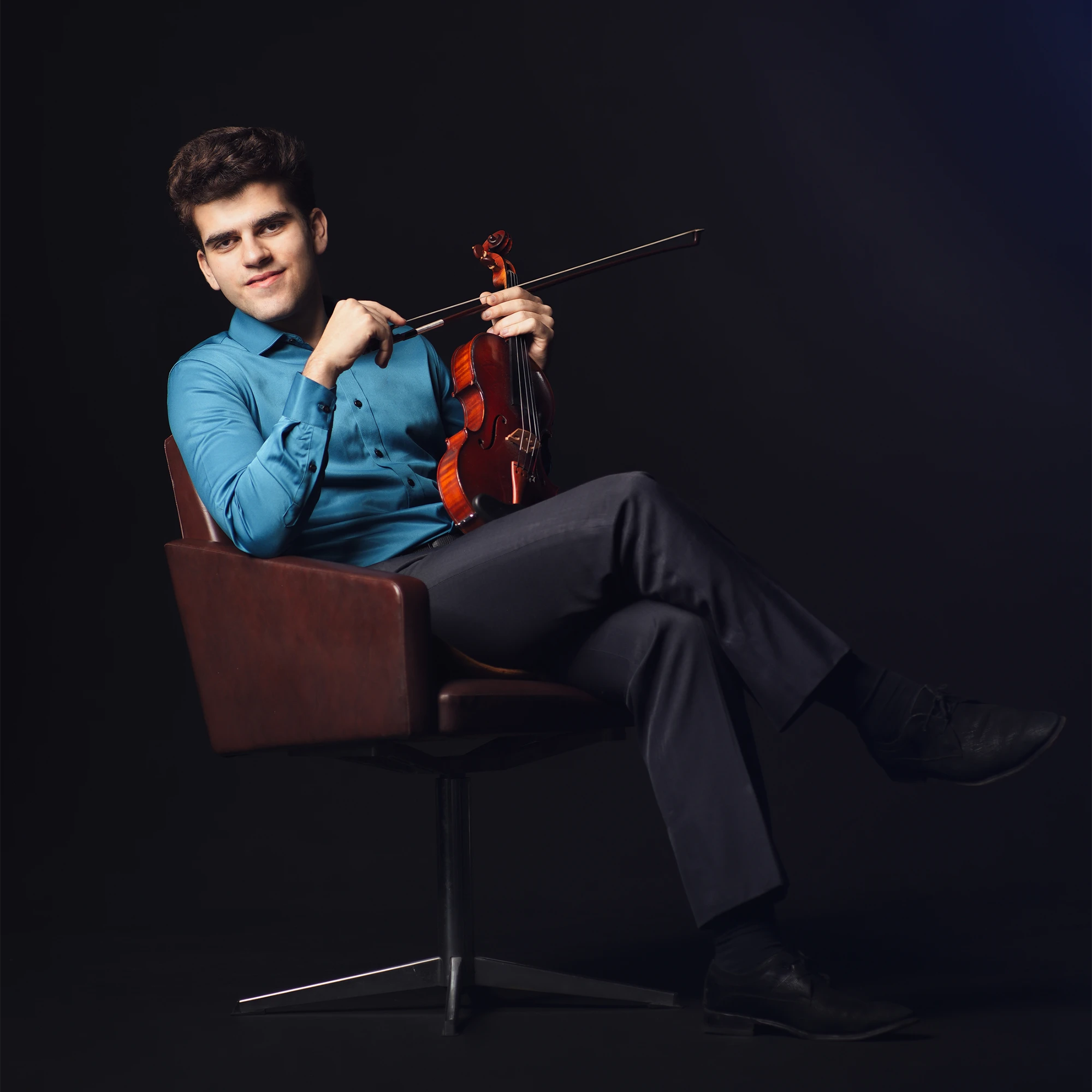 Violinist Guido Sant'Anna sitting cross-legged in a leather armchair against a dark background, wearing a bright blue button-down shirt and holding his violin and bow.