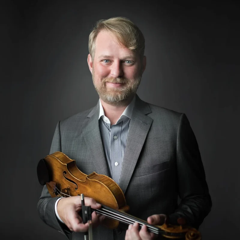 Violinist Justin Bruns smiling in a grey suit jacket and striped shirt, holding his violin and bow across his hands.