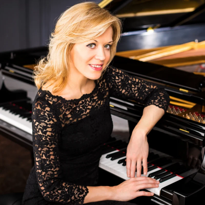Pianist Olga Kern smiling at the camera while leaning over a grand piano with her hands resting gently on the keys.