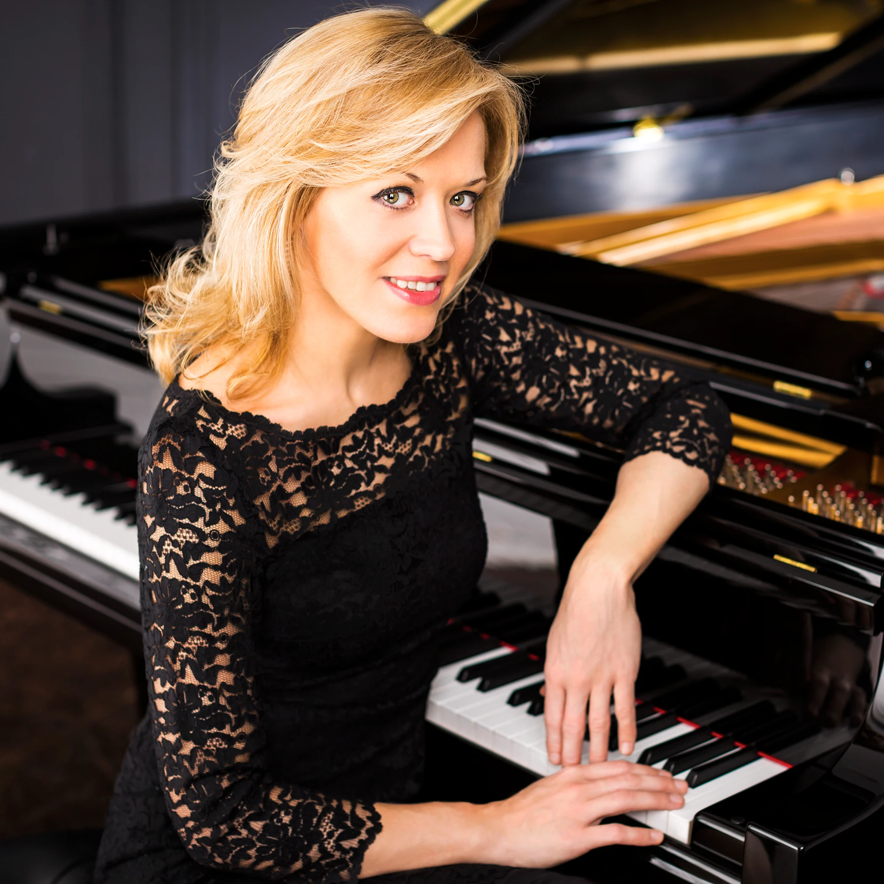 Pianist Olga Kern smiling at the camera while leaning over a grand piano with her hands resting gently on the keys.