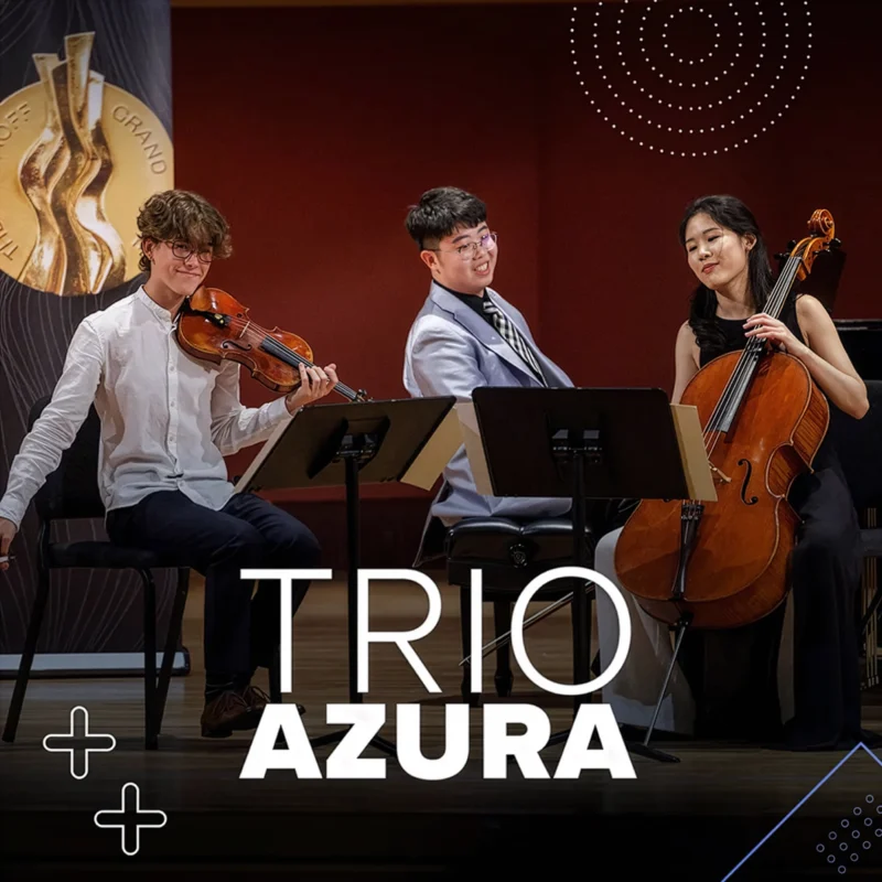 A chamber music ensemble called Trio Azura performing on stage, featuring a violinist, pianist, and cellist seated with their instruments in a concert hall with warm lighting and music stands in front of them.