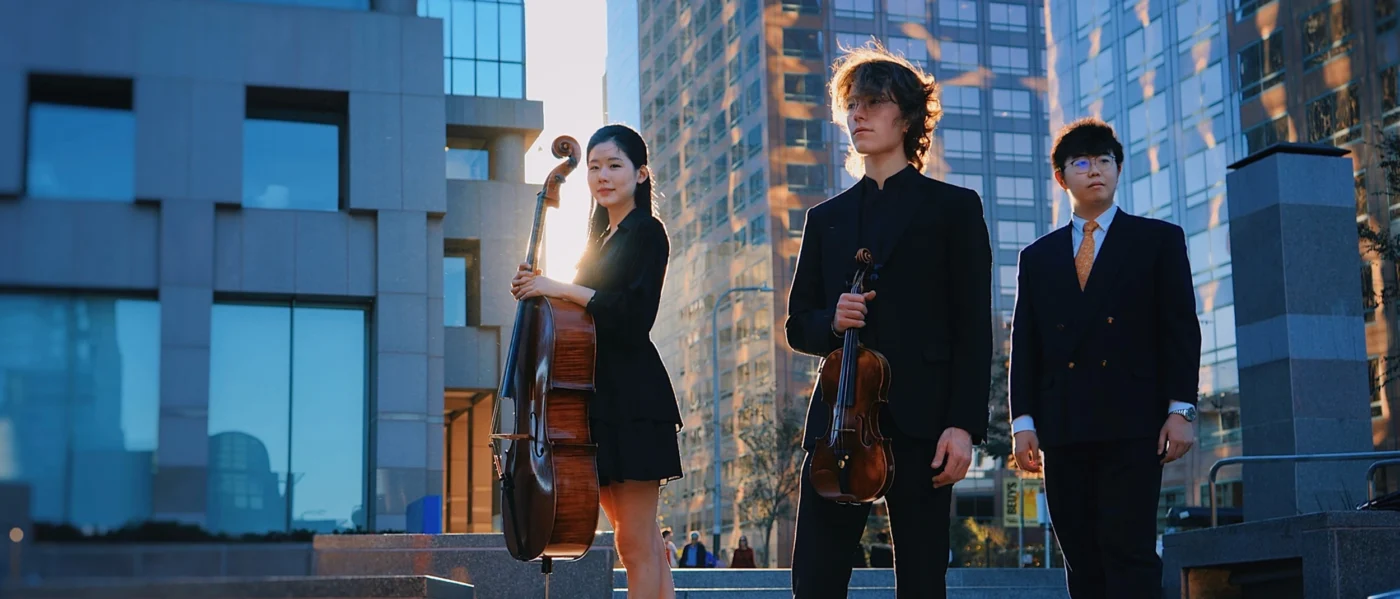 Members of Trio Azura standing outdoors in front of modern city buildings, with the cellist holding her cello, the violinist holding his violin, and the pianist standing beside them in formal black attire during a sunset photoshoot.