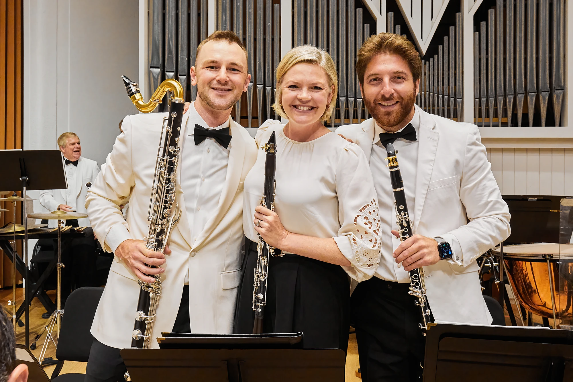 Bass clarinet and clarinet players pose together on a concert hall stage in white formalwear, surrounded by orchestra instruments and organ pipes.