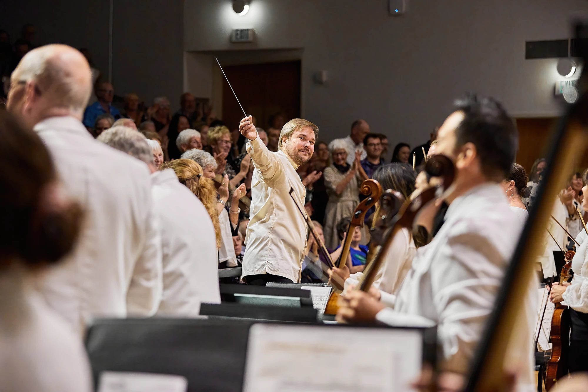 Conductor Marcelo Lehninger acknowledges the audience with raised baton on stage, surrounded by orchestra musicians as the audience applauds in a concert hall.