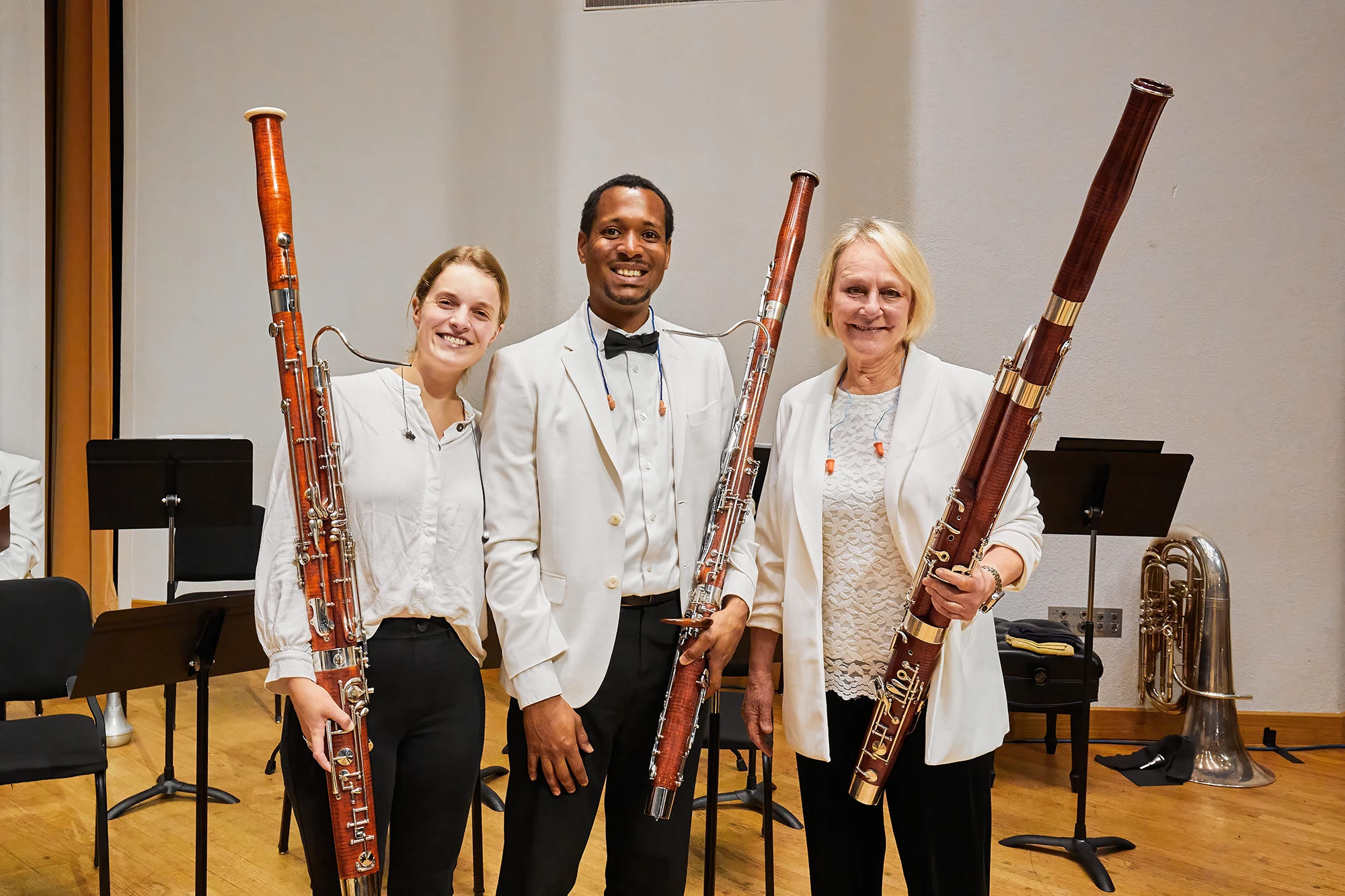 Three bassoonists in white formal concert attire smile on stage, holding bassoons in a concert hall with music stands and orchestra instruments behind them.