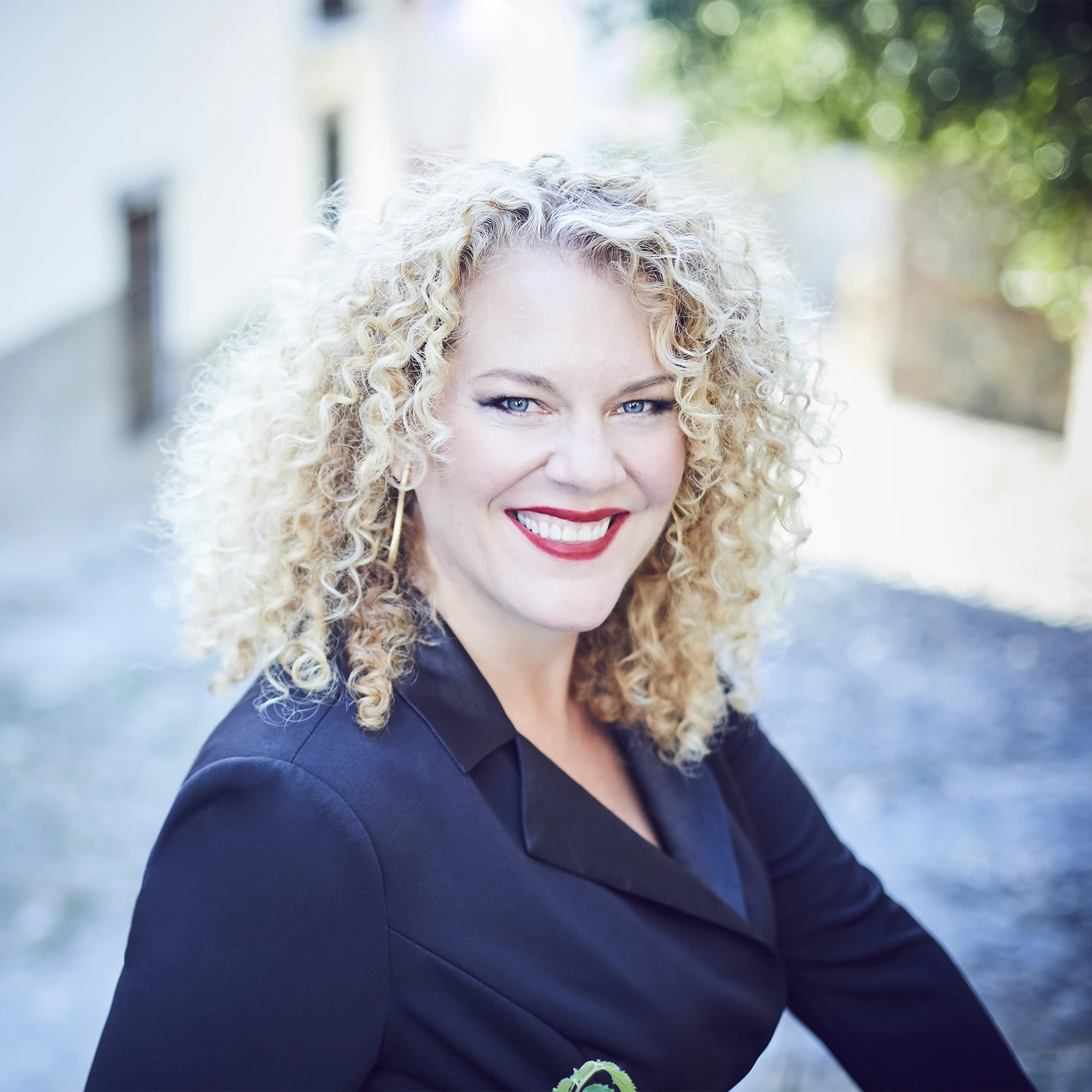 Outdoor portrait of mezzo-soprano Michelle DeYoung with curly blonde hair, smiling at the camera in a dark jacket with red lipstick.