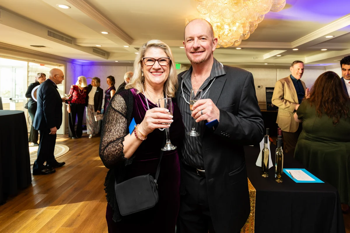 A smiling couple poses with champagne glasses. The woman wears a purple velvet dress and pearls; the man wears a dark suit over a striped shirt.