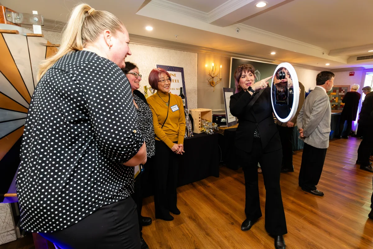 Four women stand at an interactive photo booth. One woman in a black suit points toward the ring light and camera while the others look on and smile.