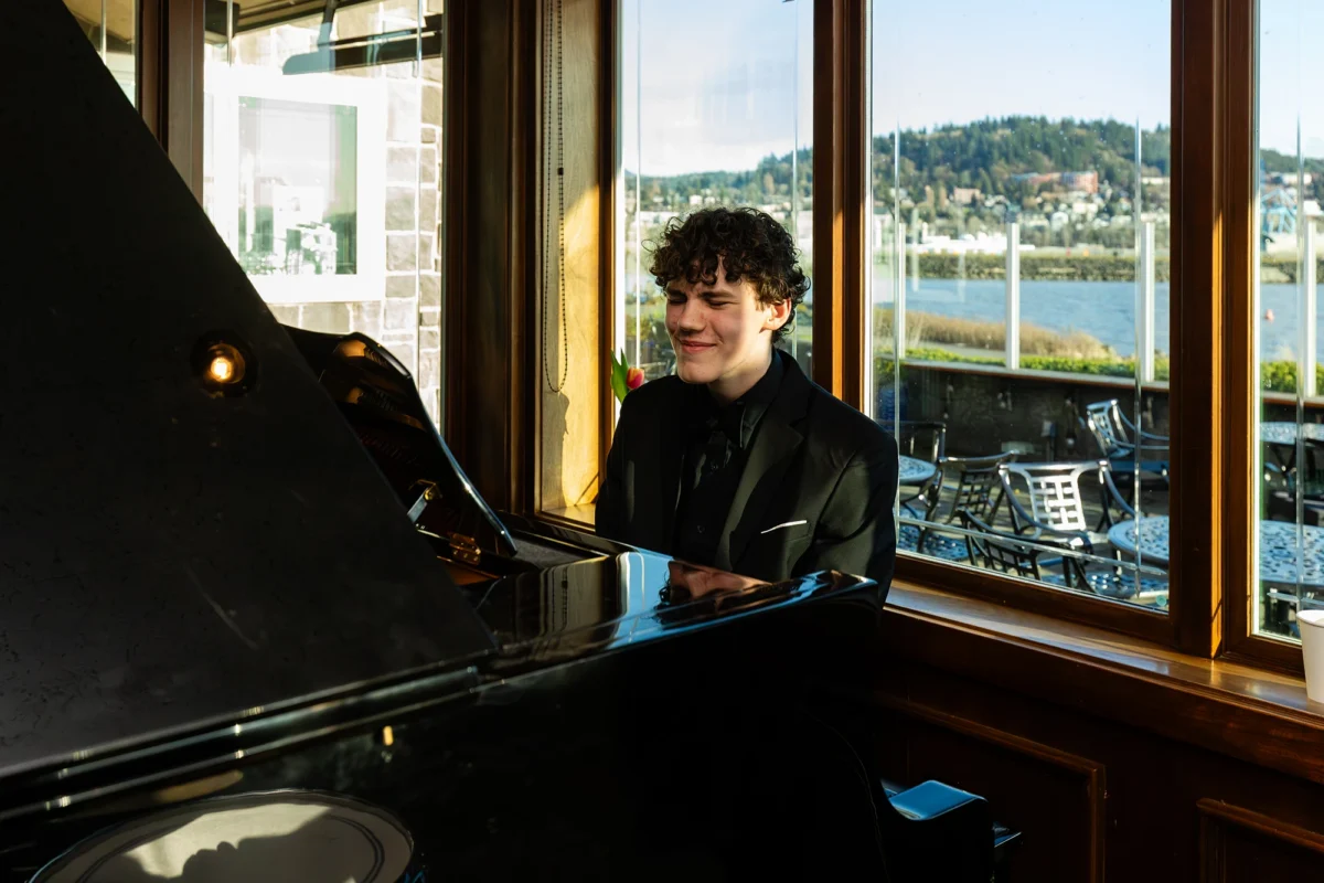 A young man with curly hair wearing a black tuxedo plays a grand piano. He is positioned in front of a large window overlooking the Bellingham waterfront and marina during the golden hour.