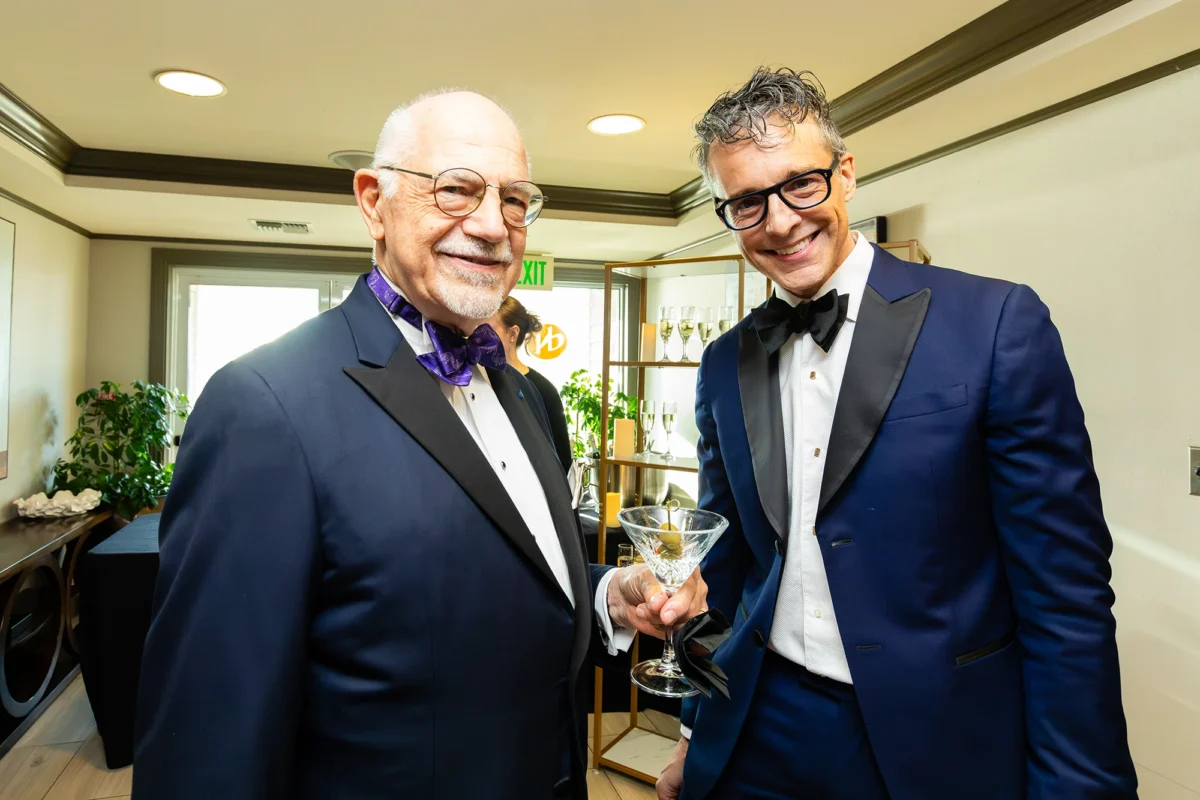 Two men smile for a photo during a cocktail hour. The man on the left is older with a white beard, wearing a dark navy suit and a vibrant purple patterned bowtie. The man on the right wears a bright blue tuxedo with a classic black bowtie and thick-rimmed glasses, holding a martini glass with an olive.