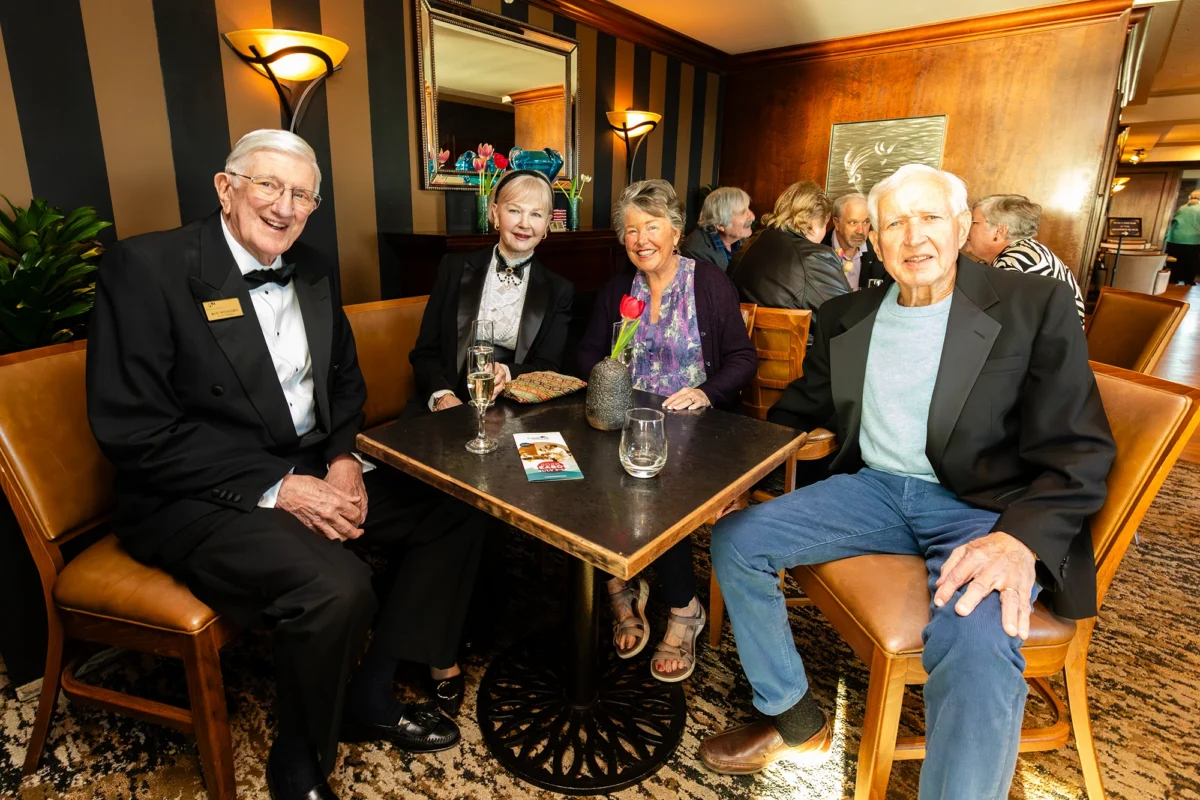 Four individuals are seated around a small, square black table in a lounge area with striped wallpaper. On the left, an older man in a tuxedo and name tag sits next to a woman in a black blazer and white lace blouse. To the right, another woman in a floral blouse sits across from a man in a light blue sweater and black blazer.