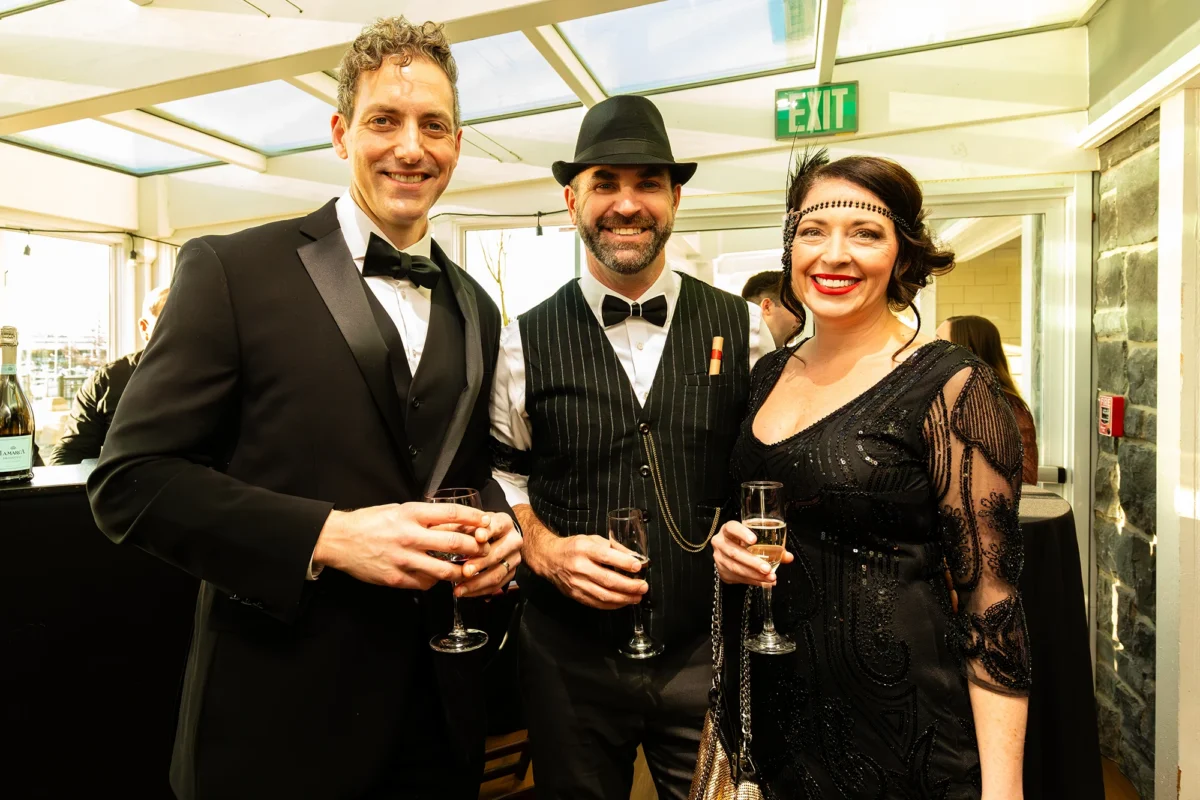 Three guests pose together, dressed in 1920s-inspired formal wear. The man on the left wears a black three-piece tuxedo. The man in the center wears a pinstriped vest, a black fedora, and a bowtie with a cigar tucked into his pocket. The woman on the right wears a black beaded flapper-style dress with a feathered headband and bright red lipstick.