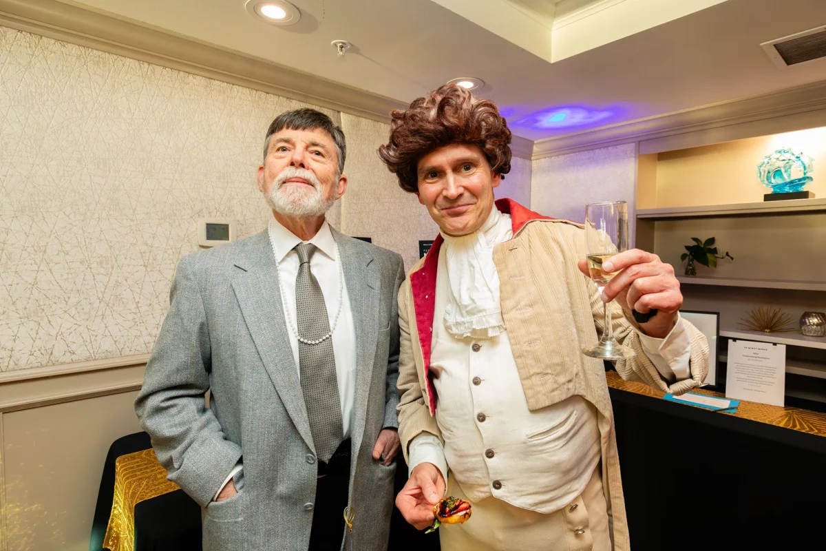 A man with a white beard in a grey tweed suit and pearl necklace stands next to a man dressed in an elaborate 18th-century style costume, featuring a powdered-style brown wig, a ruffled cravat, and a cream-colored frock coat. The man in costume holds a champagne flute and a small appetizer.