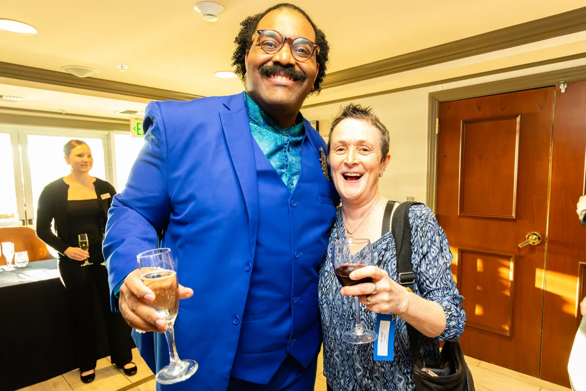 A tall man in a striking, bright blue three-piece suit with a patterned teal shirt smiles broadly alongside a woman in a blue patterned blouse. Both are holding wine glasses and appear to be laughing. A server with a tray of drinks is visible in the background.
