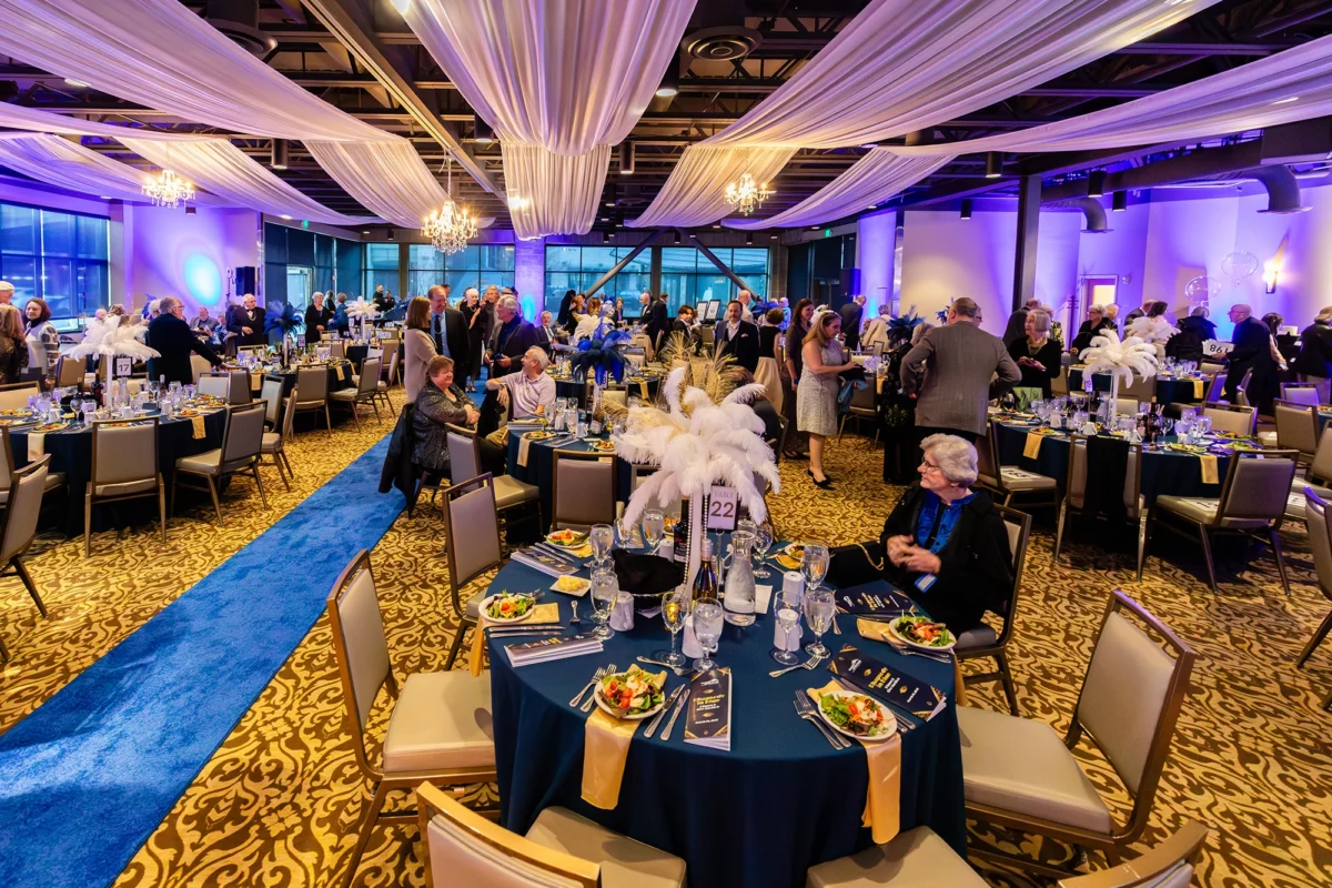 A wide view of a large ballroom prepared for a gala. The room features round tables with navy blue linens, gold napkins, and tall white feather centerpieces. A long blue carpet runner acts as a central aisle, and the ceiling is draped with elegant white fabric swags and crystal chandeliers.