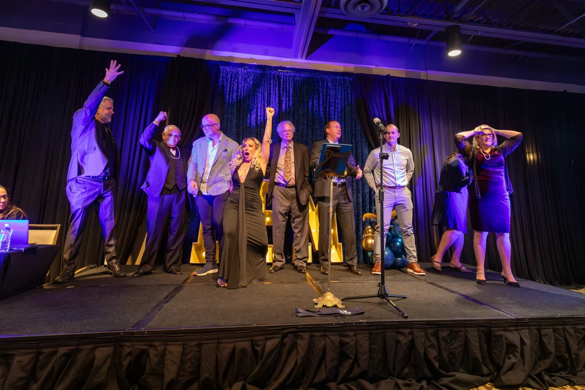 A group of eight people stand on a stage with a black curtain backdrop, many with their arms raised in celebration or cheering. They are dressed in various formal and semi-formal styles. To the left, a man in a grey suit waves, while a woman in a black dress in the center cheers enthusiastically.