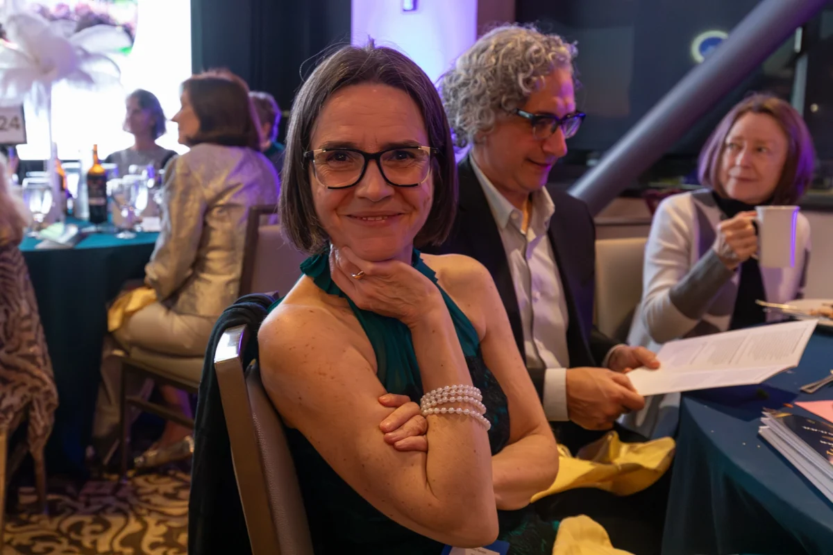 A close-up portrait of a woman with short brown hair and glasses, resting her chin on her hand and smiling at the camera. She is wearing a teal sleeveless dress and several strands of pearl bracelets. Other guests are visible in the soft-focus background at her dinner table.