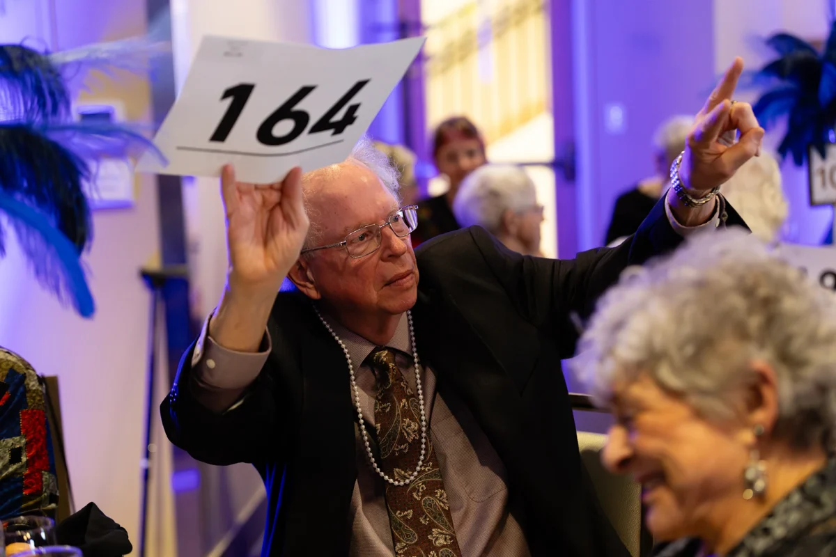 An older man wearing glasses and a pearl necklace holding up auction paddle number 164 during a live event.
