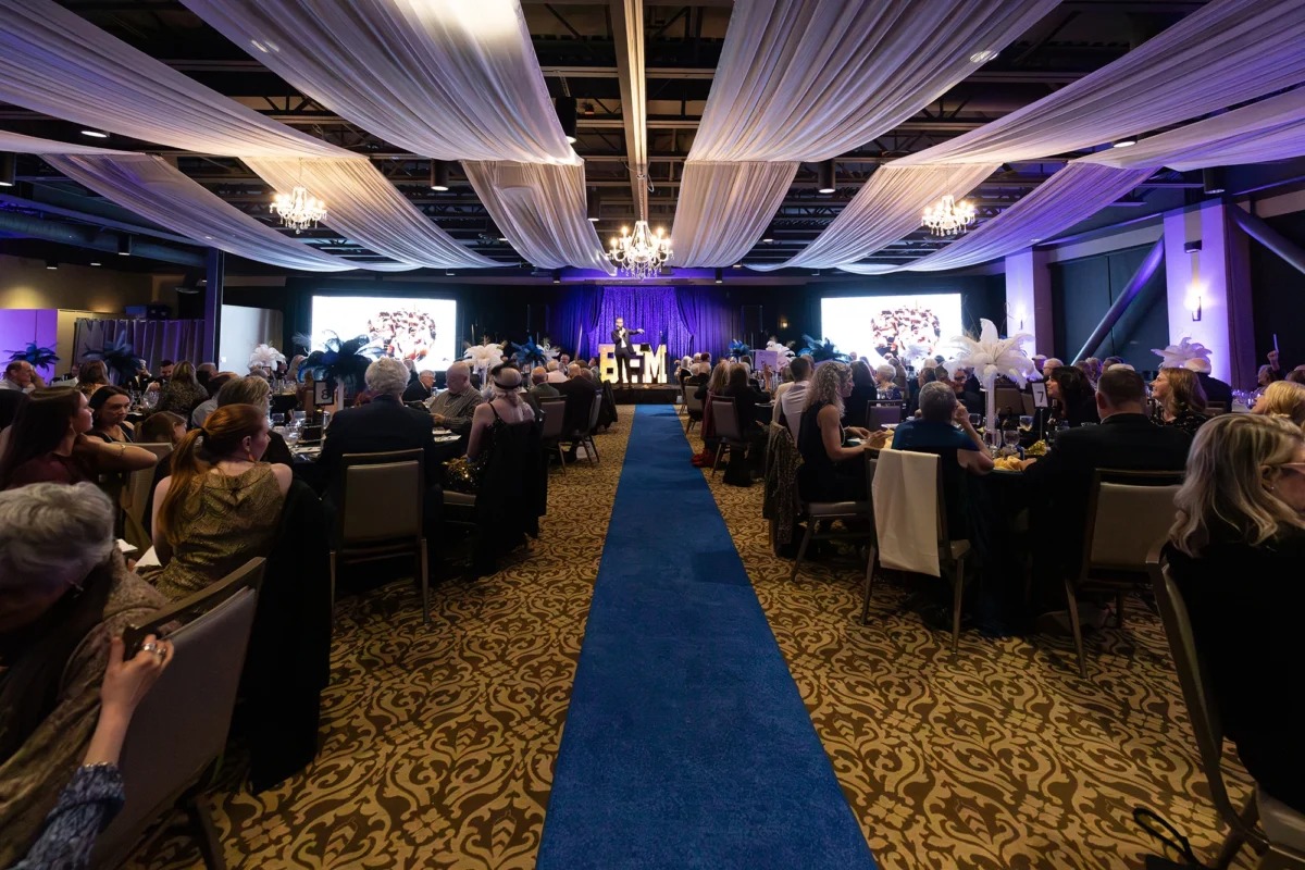 A wide-angle shot from the back of the ballroom during the event. Guests are seated at tables facing a stage where a speaker is standing in front of large "BFM" light-up letters. The blue carpet runner leads the eye toward the stage under the white draped ceiling and glowing chandeliers.