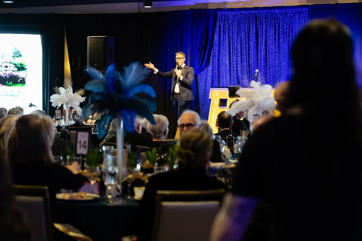 A male auctioneer in a tuxedo speaking into a microphone on a stage with blue curtains and large illuminated 'BFM' letters.