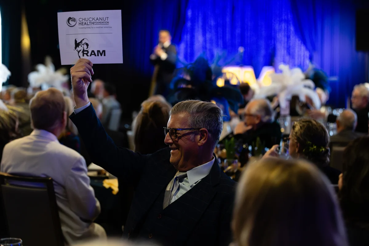 A man in a navy pinstripe suit smiling as he holds up a 'RAM' bidding card from the Chuckanut Health Foundation.