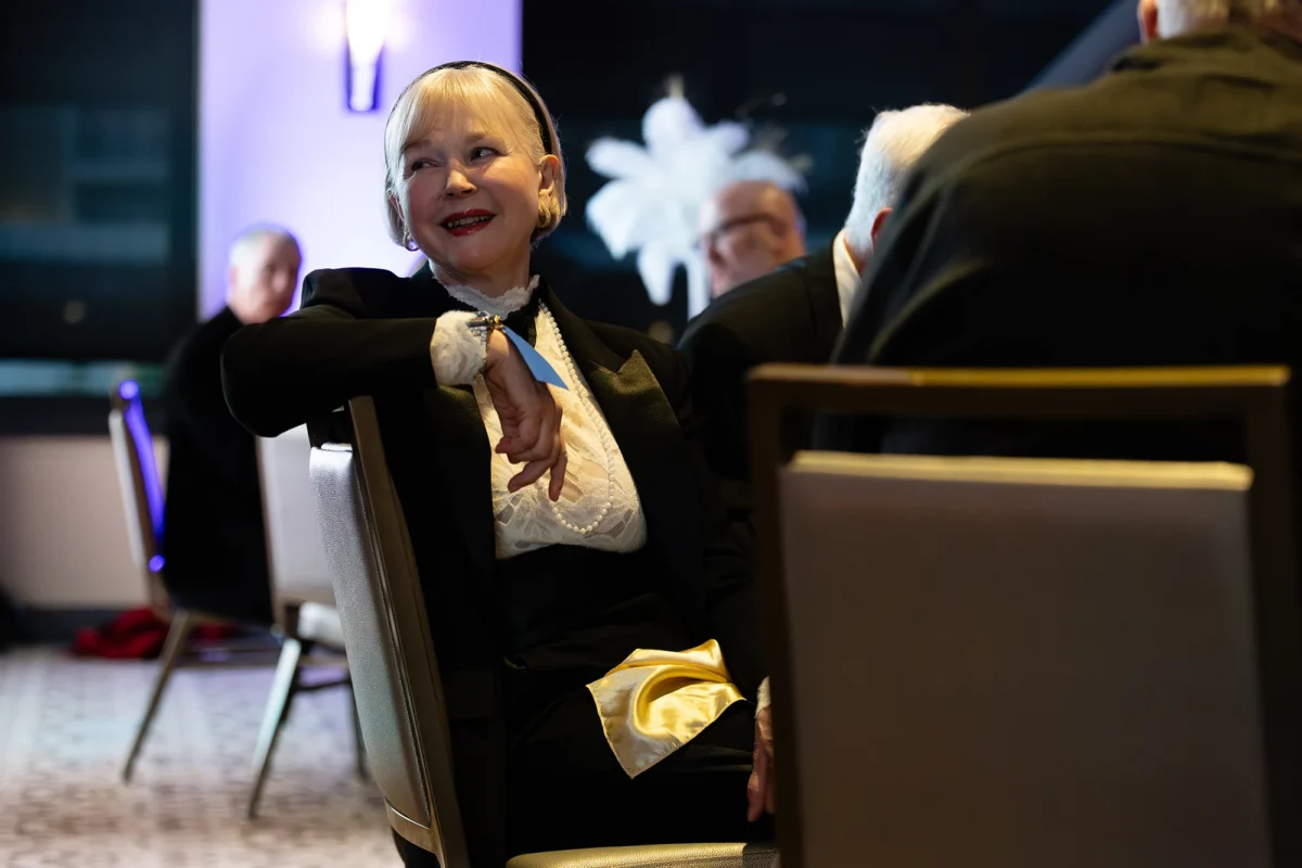 A woman in a black tuxedo jacket and white lace blouse smiling warmly while seated at a formal gala dinner.
