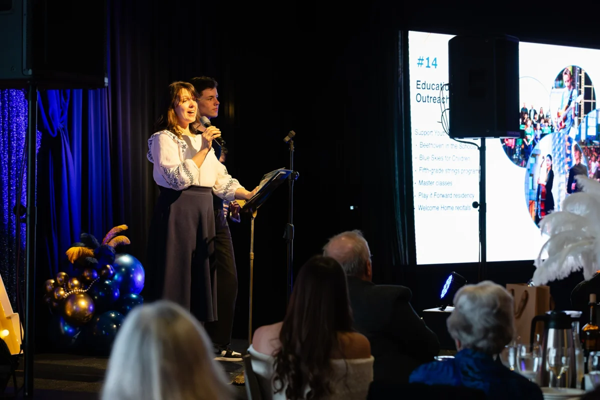 A woman in a white embroidered blouse and blue skirt speaking into a microphone on stage, with a presentation slide about education outreach in the background.
