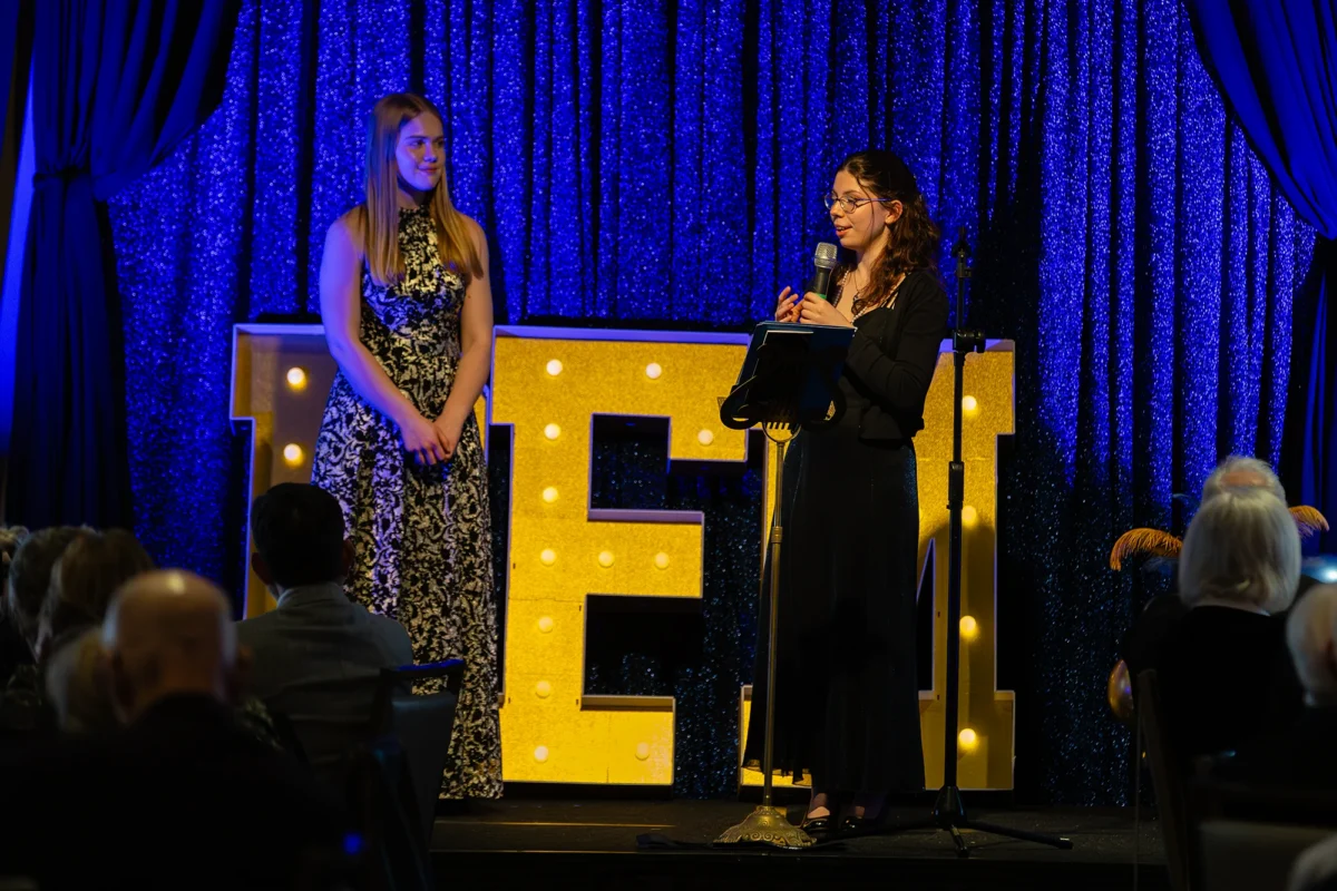 Two women standing on stage in front of a blue glitter backdrop and lit-up 'BFM' letters during a community event.