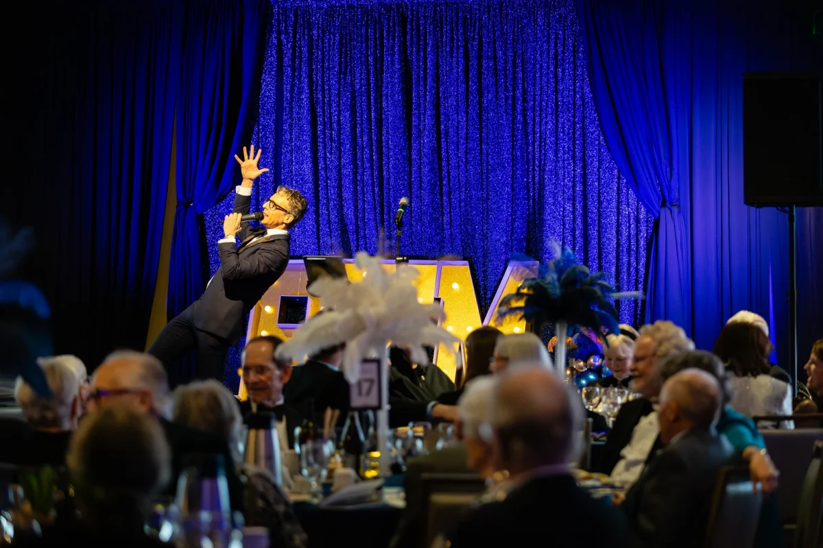 An auctioneer in a suit gesturing dramatically with one hand up while speaking into a microphone on a stage with vibrant blue lighting.
