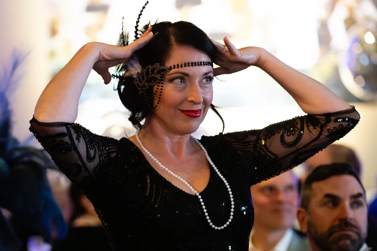A woman dressed in 1920s flapper attire adjusts a black beaded headband with feathers. She wears a black sequined dress, red lipstick, and a long pearl necklace, smiling toward the side