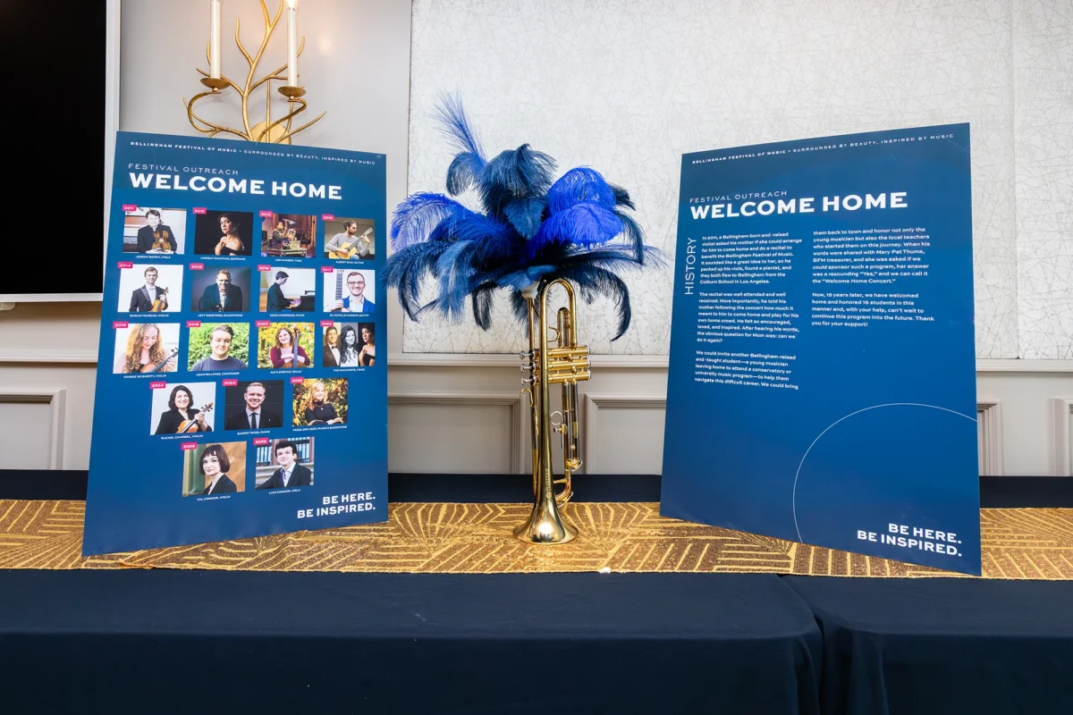 Two large blue information boards titled "Festival Outreach: Welcome Home." The boards feature photos of various musicians and a written history of the program, displayed next to a gold trumpet decorated with blue feathers.