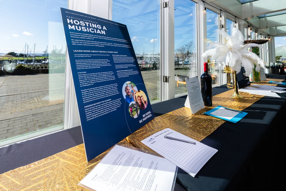 A display board titled "Hosting a Musician" sits on a table overlooking the marina. The table holds sign-up sheets, a bottle of wine, and a trumpet centerpiece with white feathers.