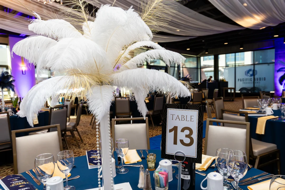 A formal dinner table (Table 13) at the gala, featuring a tall centerpiece of white ostrich feathers and pearls. The table is set with blue linens, gold napkins, and multiple wine glasses, with a "Pacific Audio Productions" van visible through the window.