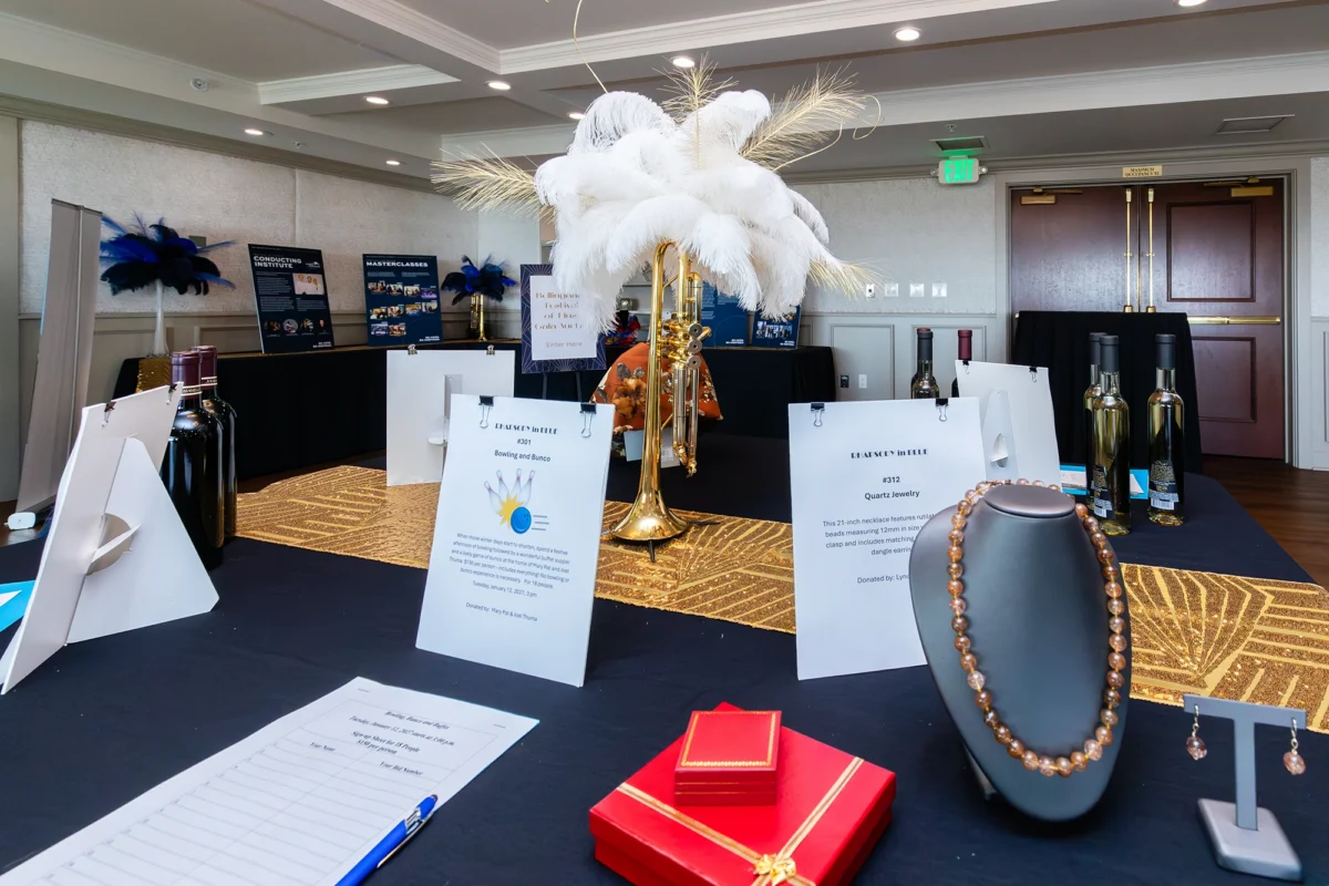 A silent auction table featuring a "Quartz Jewelry" set on a mannequin bust, a "Bowling and Busco" sign-up sheet, and several bottles of wine. A gold trumpet with white feathers serves as the centerpiece.