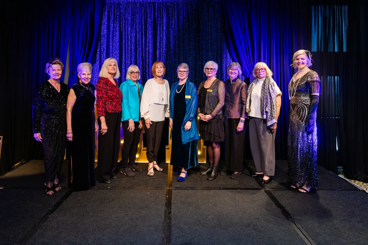 A group of ten women stand in a line on a stage in front of a blue tinsel backdrop. They are dressed in formal evening wear, ranging from colorful blouses to sequined gowns, posing for a group portrait.