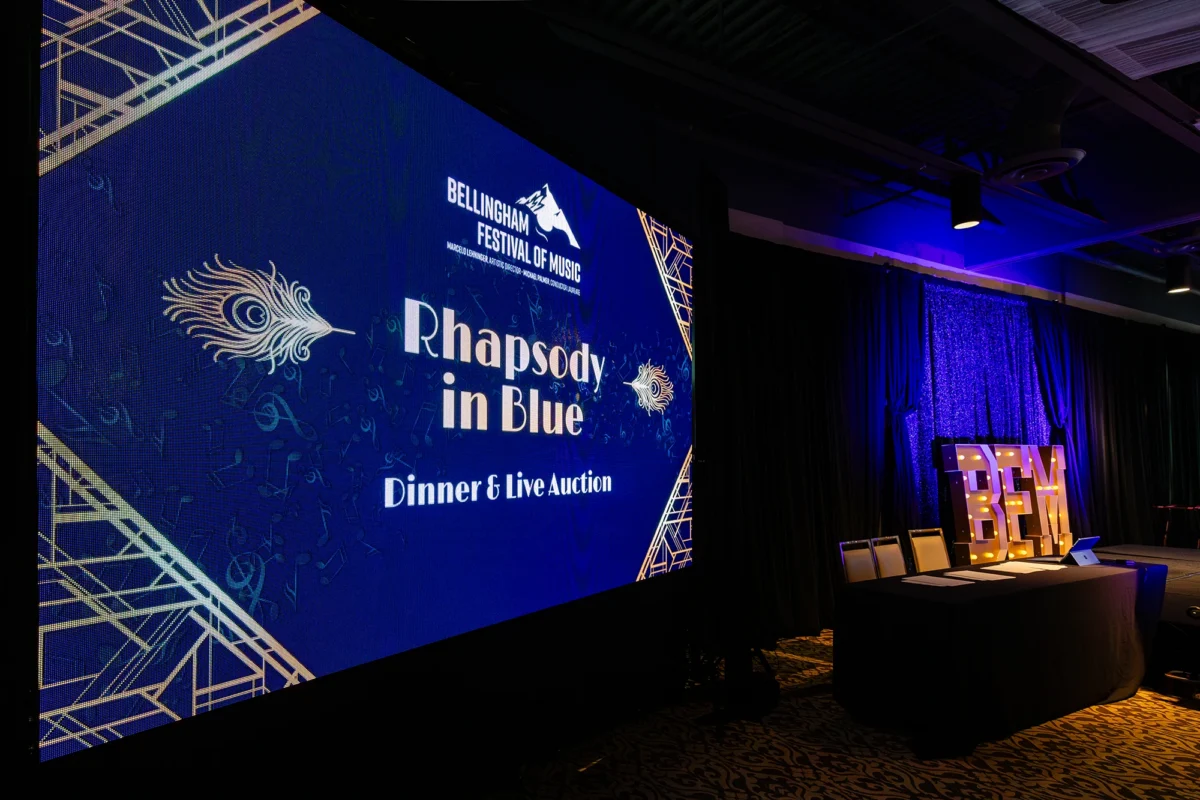 A large LED screen displays the Bellingham Festival of Music logo and the title "Rhapsody in Blue Dinner & Live Auction" in stylized Art Deco font. The screen is flanked by dark curtains and illuminated "BFM" marquee letters on a stage.