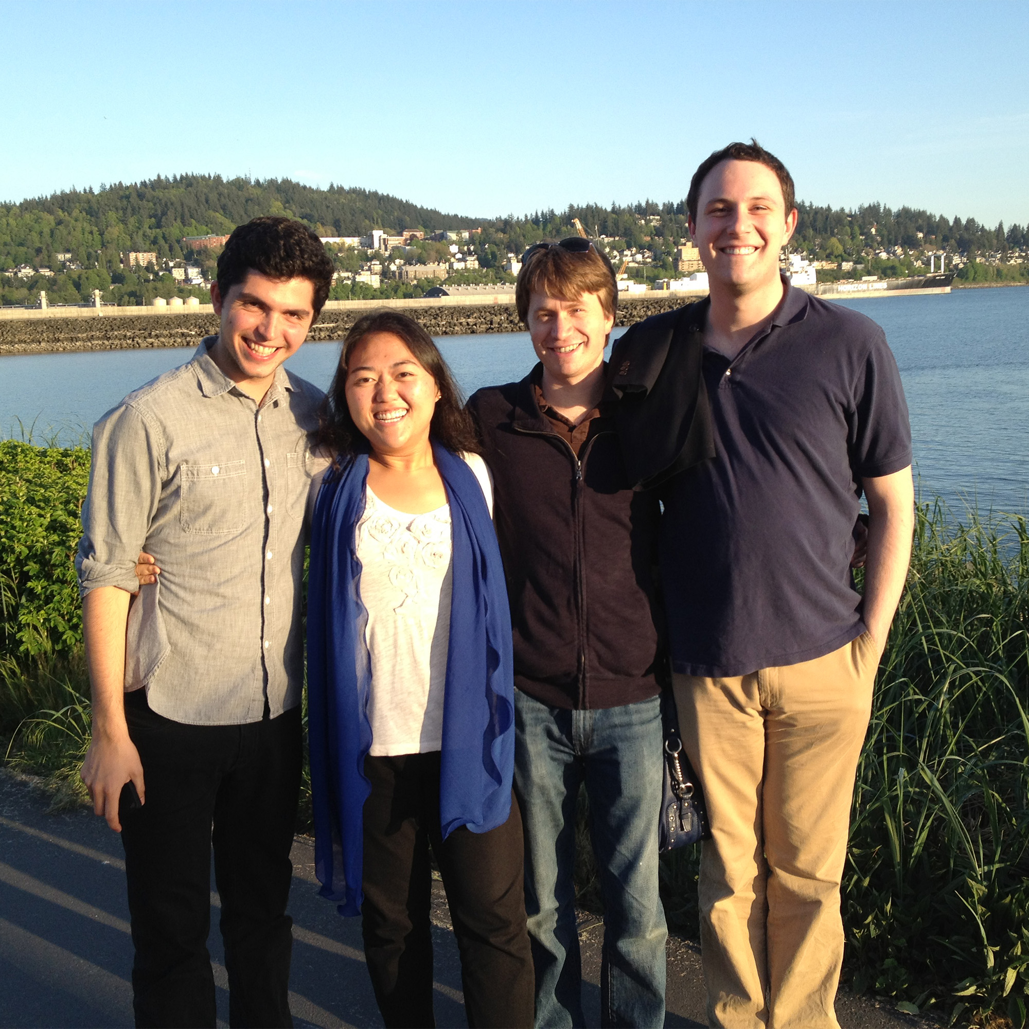 A diverse group of four young musicians smiling during the Bellingham Festival of Music, standing by the water at a Bellingham park with a view of the hillside and bay.