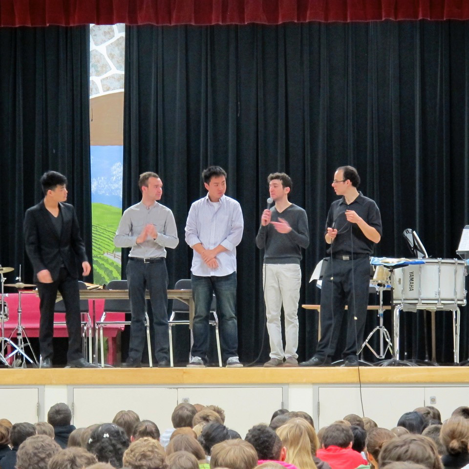 Five members of the Smoke and Mirrors Percussion Ensemble speaking to an audience during a 2013 live performance on a school stage.
