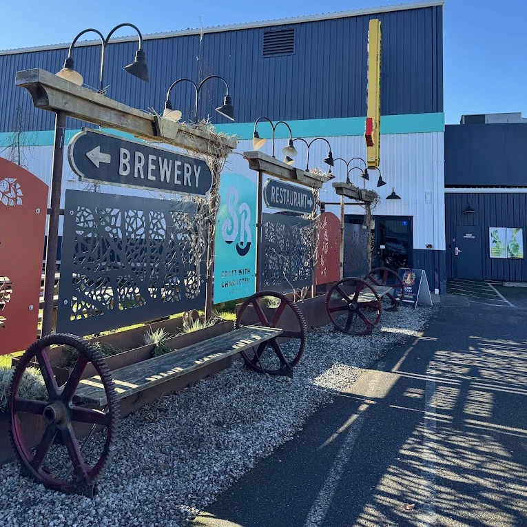 Exterior view of Stemma West brewery. In the foreground, gravel paths line a seating area with benches made of wooden planks supported by large, rusted metal gears. Signs pointing left for 'Brewery' and right for 'Restaurant' are mounted on an industrial trellis decorated with dried vines and overhead gooseneck lighting. The background shows a modern blue and white corrugated metal building under a clear blue sky.