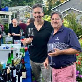BJ and Chef Jeffrey smiling at an outdoor summer event in Bellingham, holding glasses of red wine next to an outdoor bar.