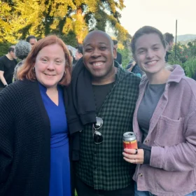 Cally, Andrew, and Julia smiling together at an outdoor evening gathering in Bellingham, holding drinks with evergreens in the background.